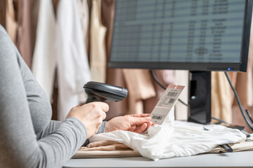 A warehouse woman employee accepts clothes using barcode scanner reading a bar code from price tag of female blouse and adds to the computer base.