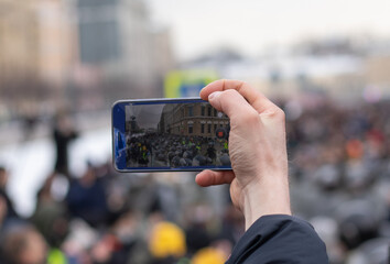 Hand with phone filming video. Police and protest demonstration on screen. Display with crowd of...