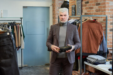 A mature man with gray hair and a sporty physique is holding a black shoe in a clothing store. A male customer with a beard wears a wool suit in a boutique.