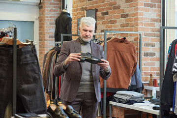 A mature man with gray hair and is choosing shoes in a clothing store. A male customer with a beard wears a wool suit in a boutique