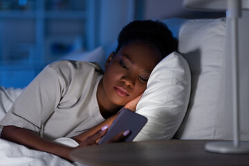 technology, internet, communication and people concept - young african american woman with smartphone lying in bed at home at night