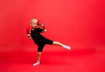 Fototapeta na wymiar a little boy boxer in red boxing gloves makes a kick on a red background with a place for text
