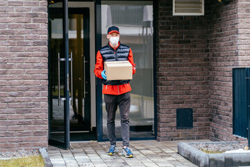Contactless delivery service concept. Courier bring parcels. Delivery boxes to door. Online order during quarantine. Man wearing protective facial mask, gloves and uniform carrying cardboard boxes.