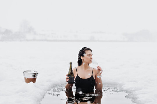 Girl with champagne in frozen lake ice hole. Woman hardening the body in cold water. Successful woman concept