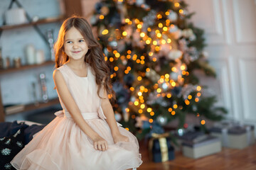 A cute brown-haired girl in a pink dress sitting near the Christmas tree. New Year