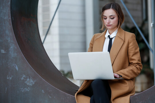 manager donna bionda con i capelli a caschetto, camicia bianca , cappotto beige e  cravatta nera, lavora al computer in esterni con espressione seria.