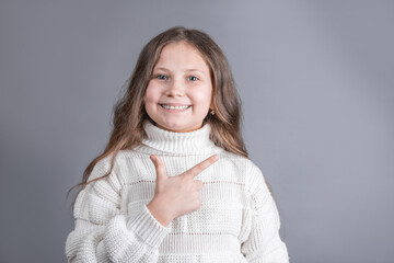 Portrait of a young attractive little girl with blond long flowing hair in a white sweater smiles and points to the side on a gray studio background. Place for text. Copy space