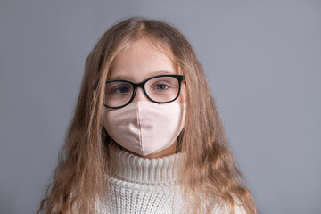 Portrait of a young attractive little girl with blond long flowing hair in a white sweater in medical protective mask on a gray studio background. Place for text. Copy space