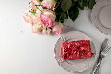 Gift with red ribbon on a plate on white background and pink roses. Top view