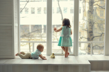 little girl in the blue dress standing at the window and looks in them, and next to her little brother lying and playing with a yellow duck