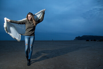 bellissima ragazza bionda con maglione verde e jeans, gioca in spiaggia con un telo bianco nell'ora...