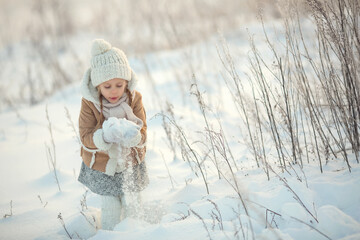 Happy little girl in white mittens and a white hat playing with snow, in a winter park, standing in the snow, throwing snow. sunset. Christmas