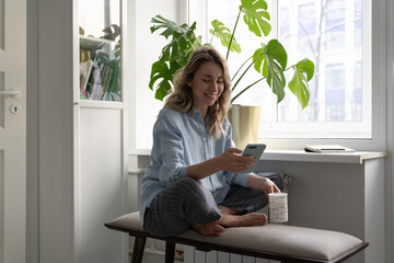 Smiling woman holding cup of coffee, using mobile smart phone, talking in video chat or social...