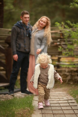 A curly little girl with blond hair in a warm waistcoat and beige trousers walks with her parents in the courtyard of a house in the village. Concept of a happy family. Village