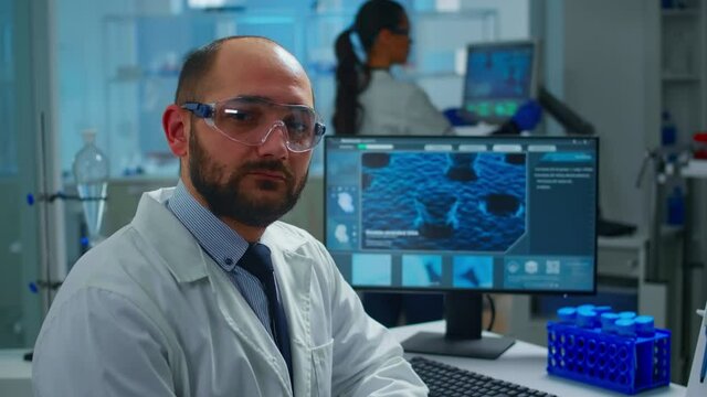 Portrait of exhausted scientist man looking at camera sitting in front of computer with DNA scan image. Chemist examining virus evolution using high tech and chemistry tools for scientific research
