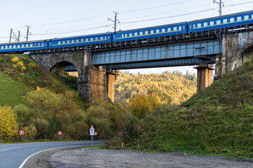The train travels along an old stone arched railway bridge over a highway in the mountainous part of the Ukrainian Carpathians. Autumn landscape - yellowed and reddened foliage, green grass.