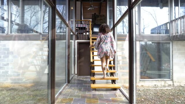 beautiful mixed race black woman wearing lingerie and robe walking up a flight of wooden stairs. she has a big curly hair afro. The sunlight shines in the window to illuminate the glass hallway