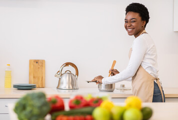 Fototapeta premium Happy African Woman Cooking Dinner In Kitchen, Smiling To Camera