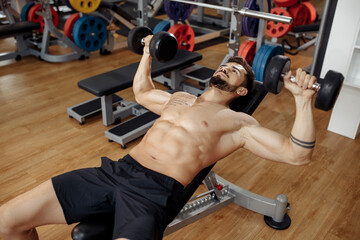 Muscular athletic young man bodybuilder fitness model exercise chest with dumbbell on the bench in gym. Top view.