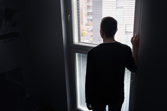 Lonely man looking out the window. Solitude, isolation and loneliness concept. Sad upset person with regret, shame or stress in moody dramatic back view silhouette. Dark room with shadow at home.