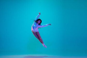 Flying. Young and graceful ballet dancer isolated on blue studio background in neon light. Art, motion, action, flexibility, inspiration concept. Flexible caucasian ballet dancer, moves in glow.