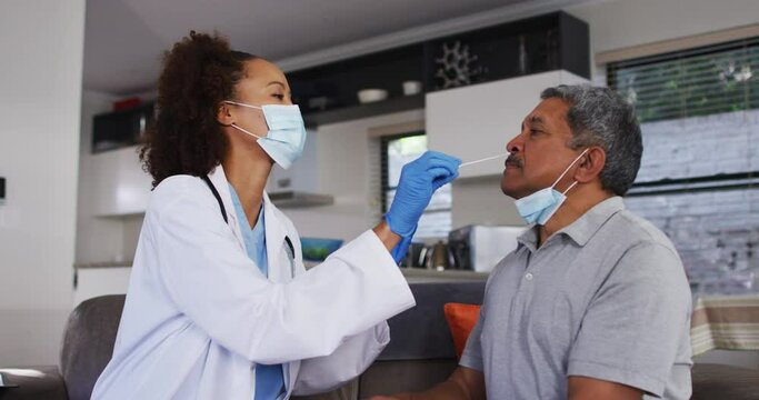 Mixed race female doctor wearing mask doing swab test on senior man at home