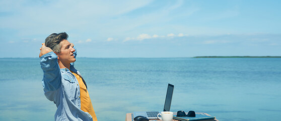 Panoramic image relaxed man freelancer enjoying morning by sea at beach. Freedom, remote job, freelance work concept, dream office job workplace