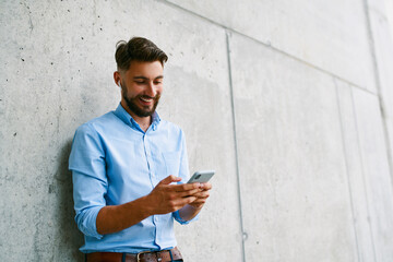 Young man texting with mobile phone standing in the city against concrete wall
