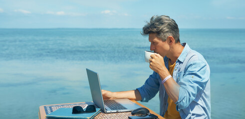 Pensive man freelancer working on laptop computer at beach by blue sea, drinking coffee. Concept remote work, freelance