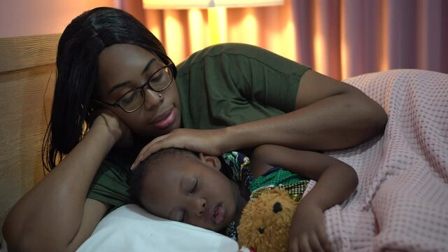 african american mother singing lullaby for little daughter before going to Sleep on bed at home. black mom story telling to girl in bedroom. sleeping child in parent embrace . kid hug Teddy bear