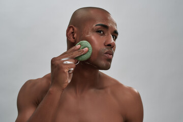 Portrait of handsome african american young man enjoying skincare procedure, using cleansing sponge while washing his face isolated over gray background