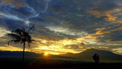 Sonnenaufgang in tropischer Landschaft beim Vulkan Mt. Gede mit gelbem Schein vor dunklen Wolken