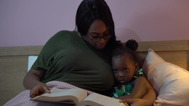 african american mother reading a book to her little daughter in bed before going to sleep at home. black mom story telling to girl in bedroom. sleepy child in parent embrace . kid with bedtime