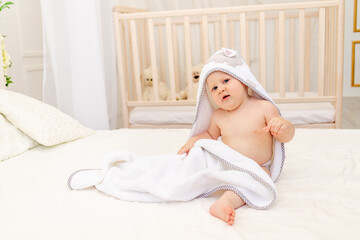 a baby in a towel sits on the bed after bathing and smiles