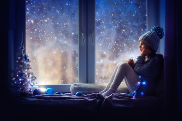 A girl in a blue hat sits on the windowsill and looks at the snow. Next to her is a Christmas tree, a garland is burning, there is a candle