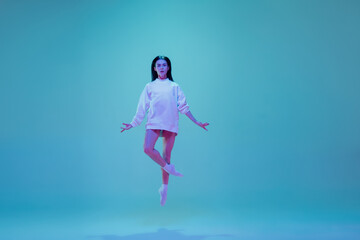 In flight. Young and graceful ballet dancer isolated on blue studio background in neon light. Art, motion, action, flexibility, inspiration concept. Flexible caucasian ballet dancer, moves in glow.