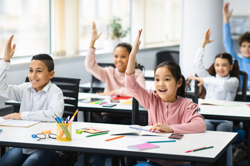 Diverse small schoolchildren raising hands at classroom