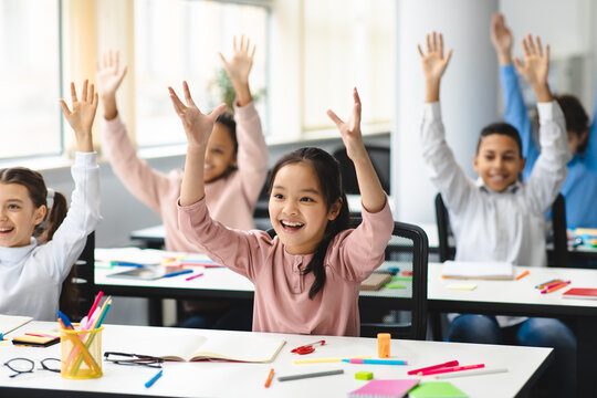 Happy diverse small schoolchildren raising hands at classroom