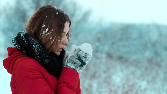 A gentle caucasian girl is standing in the snow and with a slight smile on her face warms her hands in the snowfall, blows mouth on her gloves  and snowflakes stick to her brown hair. Side view