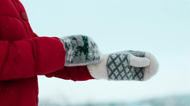 In snowfall, girl in red down jacket and with black knitted scarf around his neck wears white gloves made of animal fur with winter pattern with deer and abstract diamonds on background of cloudy sky