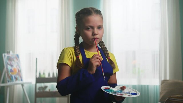 Little girl with two braids in blue apron, artist, is standing studio, reflecting on some creative idea, looking on camera and holding painting tools, Zoom in, Slow motion.