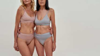 Cropped shot of two caucasian women in underwear looking at camera while posing together over light background. Warmth in relation, mature beauty concept