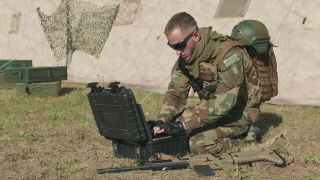 Full shot of intelligence army officer sitting on floor at military base outdoors, transmitting data via laptop