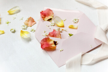 Rosaner Briefumschlag mit weißem Seidenband und Frühlingsblüten als Flatlay auf einem weißen Untergrund