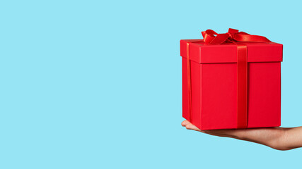 Hand holding red gift box with ribbon bow isolated on light blue background. Holiday concept. Copy space.