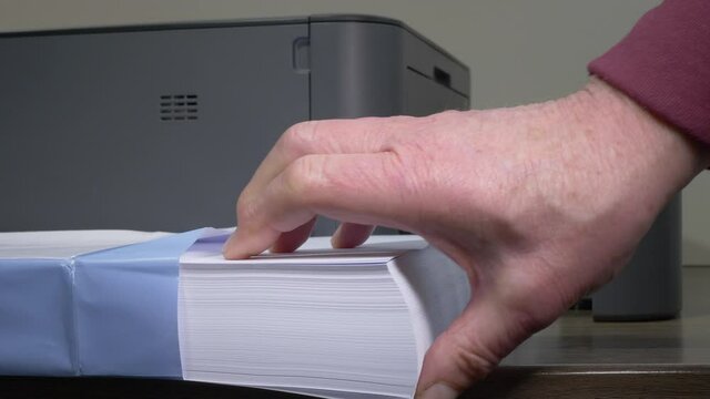 Closeup POV shot of a man&rsquo;s hand pulling out a ream of white paper from a new pack, in front of an open printer tray on a desk, then using fingers and thumb to loosen up the paper.
