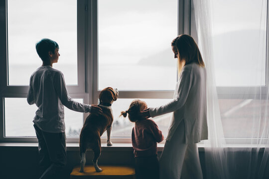 Woman with daughter, son and dog standing near the big window at the modern apartment.