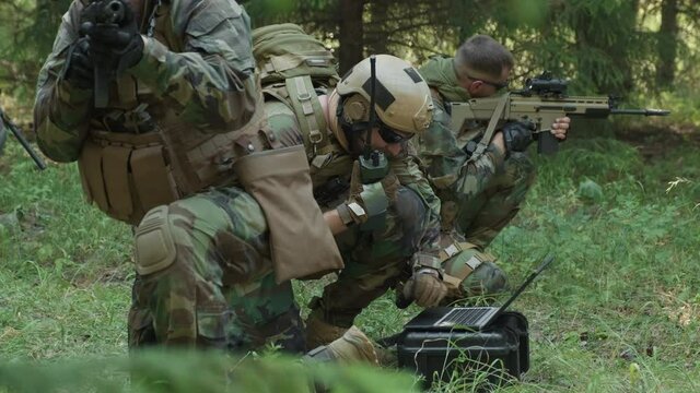 Medium shot of squad of special forces soldiers on military operation in jungle, commander using radio while transmitting data on laptop, and others covering him with rifles