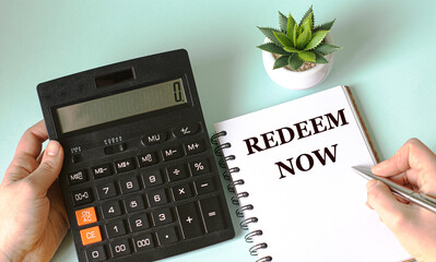 A woman's hand writes in a notebook the words REDEEM NOW on the background of a calculator