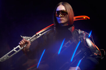 Serious military woman in stylish dark clothing and with implant on her shoulder poses in dark background. Attractive female soldier holding a glowing sword on her shoulder and a rifle.